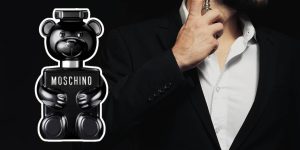 O perfume Toy Boy, com seu frasco em formato de ursinho de pelúcia metalizado, com um homem aplicando um perfume no pescoço na direita