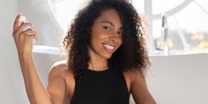 Mulher negra sorri para a câmera enquanto utiliza um spray em cima mesma