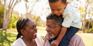 Casal de idosos sorrindo; o homem carrega uma criança, também sorridente, nos ombros