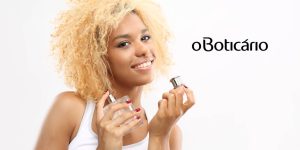 Mulher negra de cabelos loiros segura um frasco de perfume sorrindo; ao seu lado a logo do Boticário